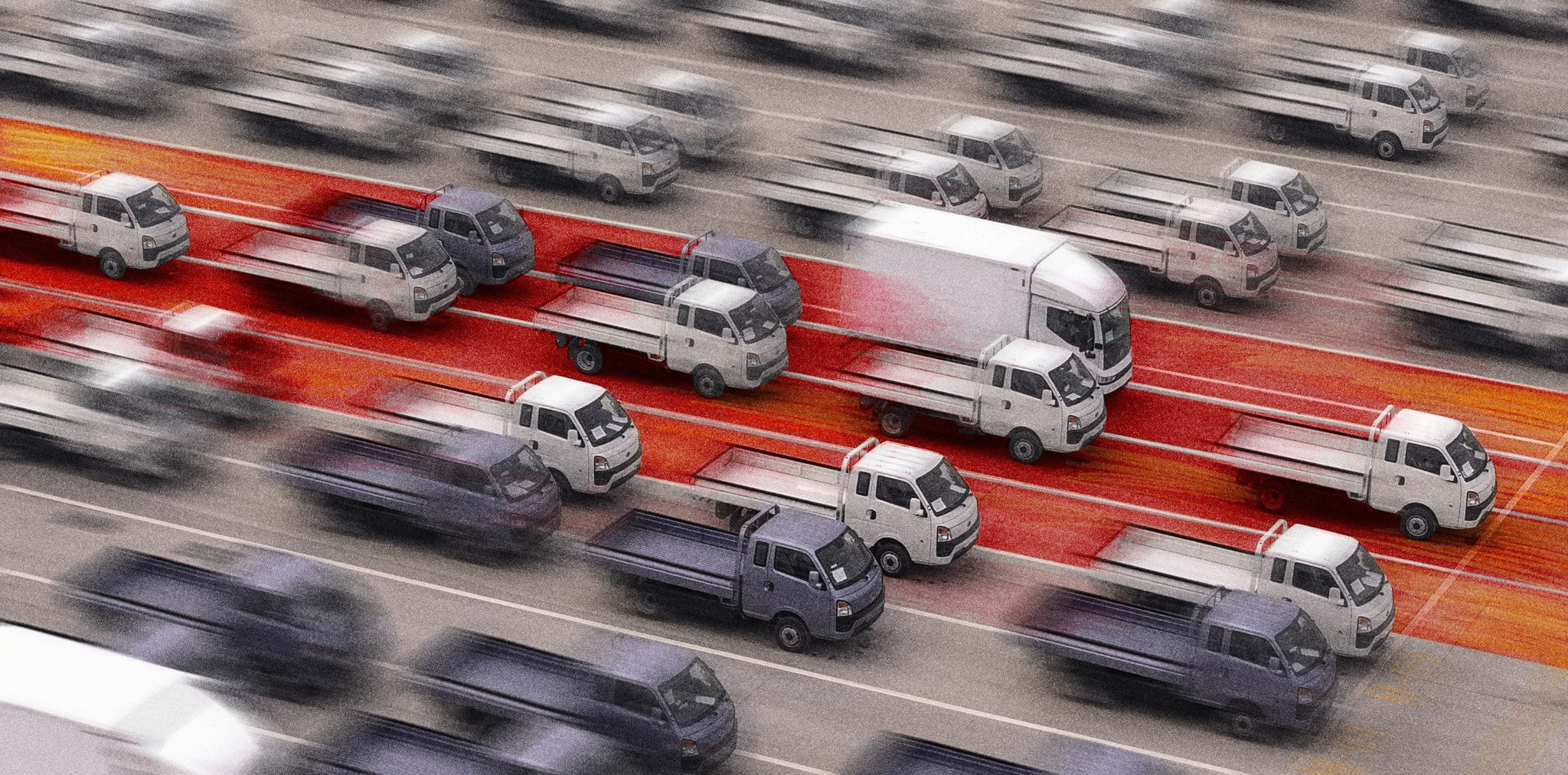 An aerial view of a busy road with a blurred motion effect, showcasing a traffic jam of small trucks and vans in different shades of gray and white, with a distinctive red lane in the center.